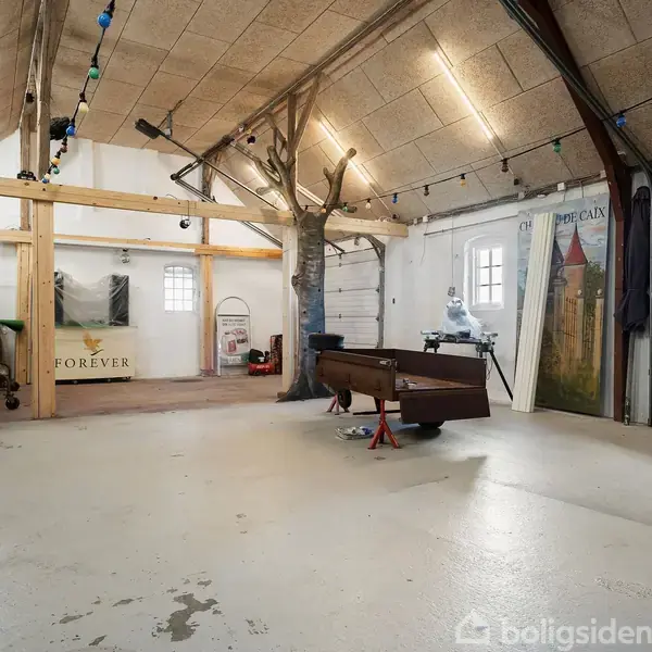 En stor garage med et træstativ i midten, dekoreret med lyskæder. Et bord står placeret nær en dør. Væggen har et maleri og en plakat med teksten "FOREVER".