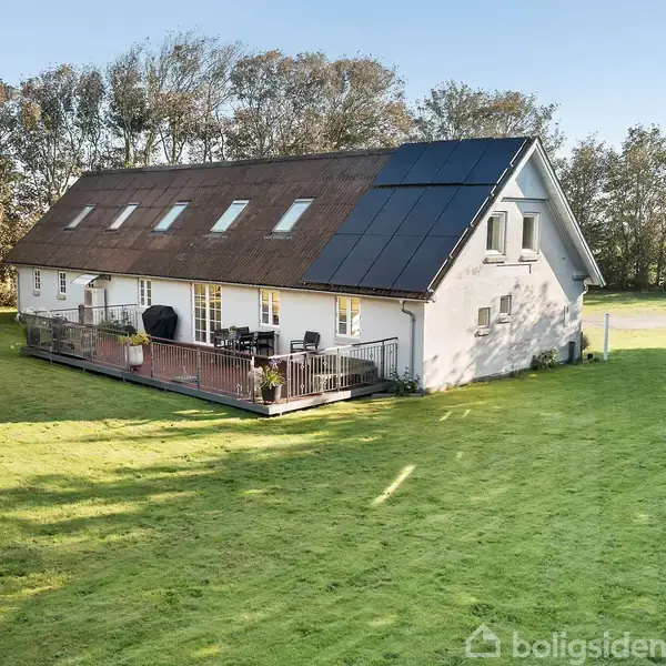Et moderne hus med solpaneler på taget står på en velplejet græsplæne. En træterrasse med havemøbler er tilknyttet. Træer omgiver området, der ser roligt og landligt ud.