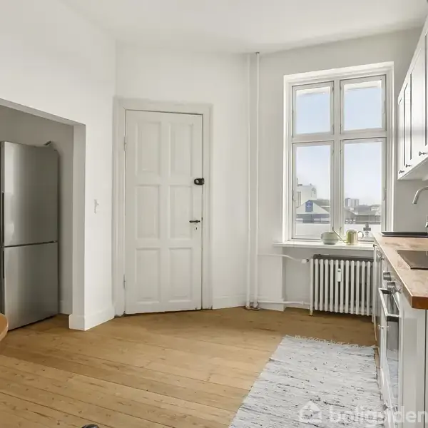 Et køkken med hvidt interiør og træbordplade. Et rundt bord med stol og keramikkrus. Køleskab ved siden af en hvid dør. Sollys strømmer ind gennem et stort vindue.