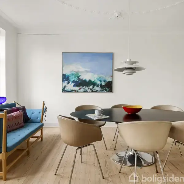 En spisebordssæt med seks beige stole står rundt et ovalt træbord. En blå sofa er placeret ved siden af et vindue. På væggen hænger et maleri.