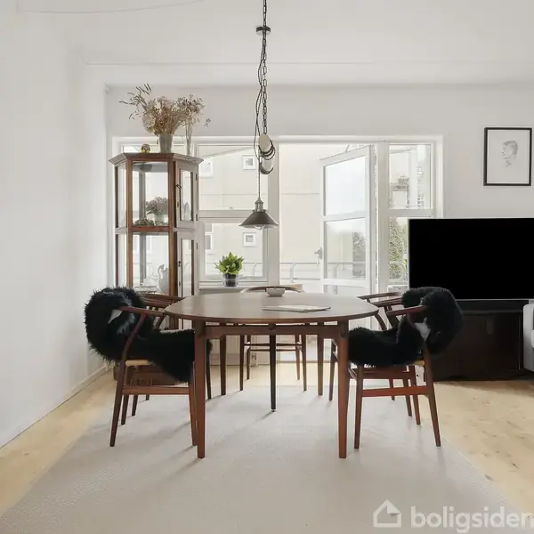 Rundt spisebord med sorte stole, står i en lys stue med åbne vinduer og hængende lamper. Et gråt sofaarrangement med puder ses ved siden af et fjernsyn.