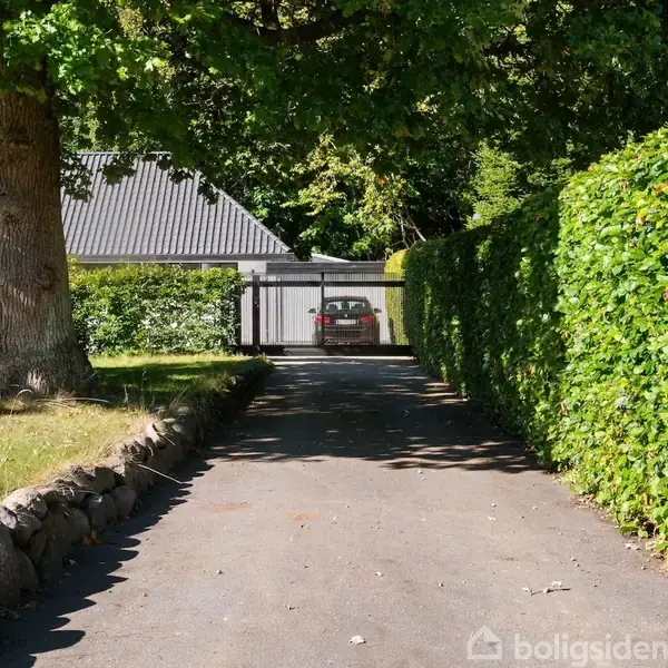 En bil står parkeret foran en garage, omgivet af grønne hække og en stor skyggefuld træsti. Stien er afgrænset af en lav stenmur, der fører op til huset.