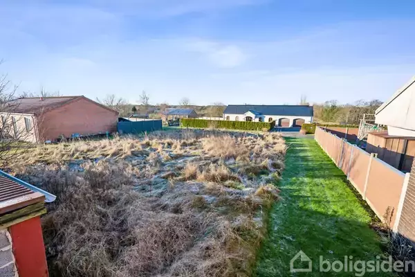 En tom grund med græs og buske ligger mellem to huse. I baggrunden ses et større, moderne hus og en blå himmel. Grusstier ses på begge sider af grunden.