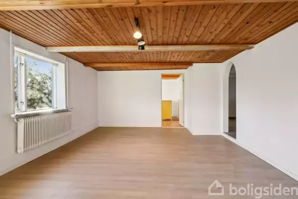 Et rum med trægulv og træloft, tomt og med lysindfald fra et stort vindue. I baggrunden ses åbninger til andre rum, omgivet af hvide vægge.