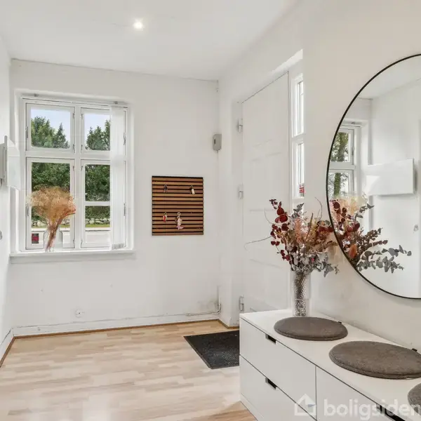 Spejl hænger på væggen over en hvid kommode med blomsterdekoration. Jakke hænger på knage. To vinduer slipper lys ind i det lyse rum.