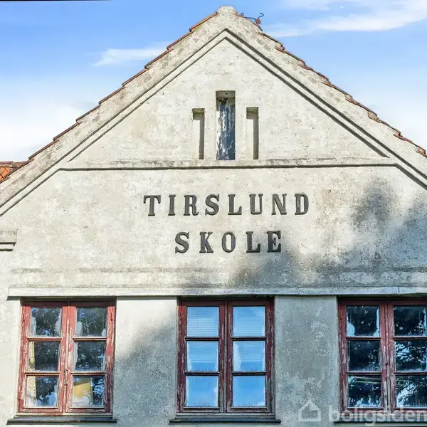 Bygning med firkantede vinduer i en murfacade. Teksten "TIRSLUND SKOLE" står skrevet på facaden. Røde teglsten udgør taget under en blå himmel.