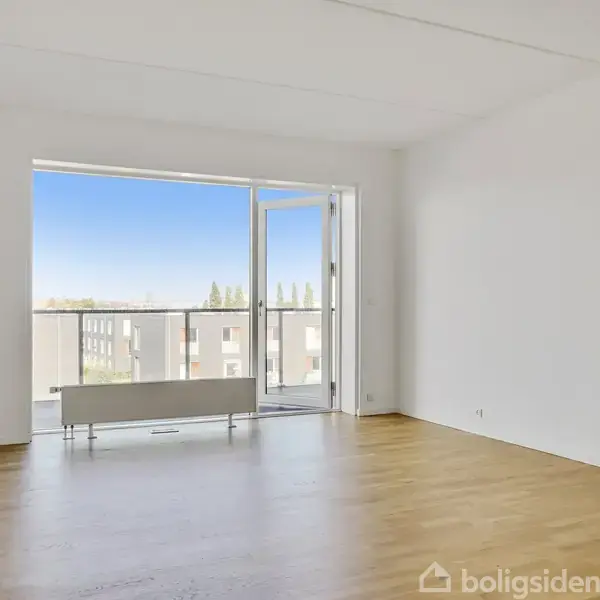 Et tomt værelse med trægulv har en åben altandør, der viser en balkon og en udsigt over bygninger i det fjerne. Væggene er hvide, og der er lyst udefra.