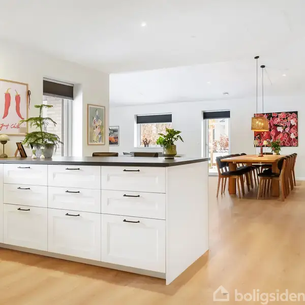 Et lyst køkken med hvidt skab og sorte håndtag, indeholder planter og vægkunst. Det åbner op til en rummelig spisestue med trægulv, spisebord, stole og dekorative lamper.