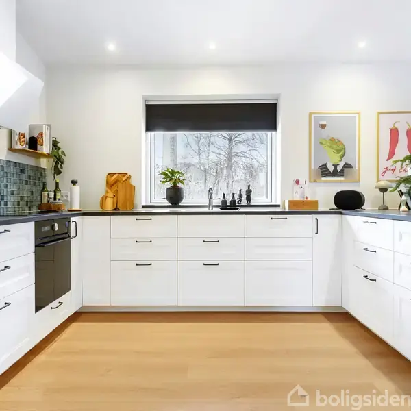 Hvidt køkken med moderne indretning, integrerede skabe og skuffer, sort bordplade. Grønne planter og vægkunst pynter. Vindue midt i rummet giver udsigt til snedækket landskab.