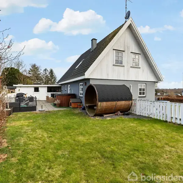 Et hvidt hus med spidst tag står ved siden af en stor tønde i en grøn have. Haven er omkranset af hække og hvidt stakit, og himlen er blå med skyer.