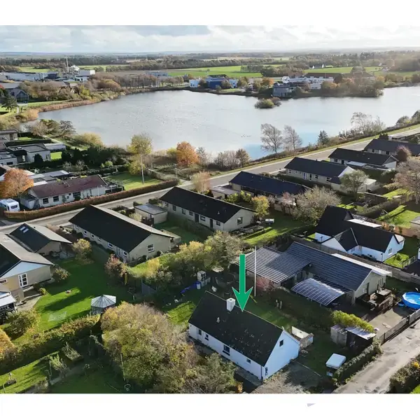 Et parcelhus markeret med en grøn pil, set fra luften, ligger i en række af huse i et parcelhuskvarter. I baggrunden ses en stor sø omgivet af landskab.