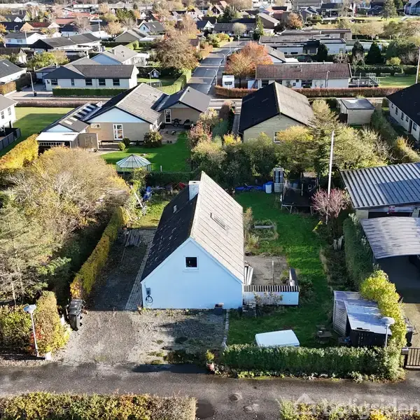 Et hvidt hus med skrått tag står i en boligkvarter. Omgivet af grønne haver og en grusindkørsel. Flere huse ses i baggrunden i et velordnet nabolag.