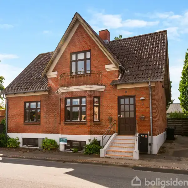 Et rødt murstenshus med spidst tag står stille i et beboelseskvarter. Huset har hvide detaljer rundt om vinduerne og en central hoveddør med trapper op til den.
