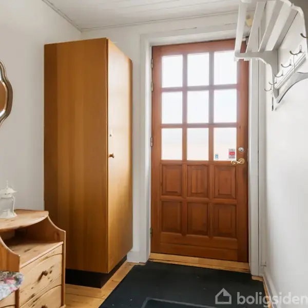 Døren med glasruder står stille i en entré. Ved siden af hænger knagerækker og overfladen er dekoreret med et gammelt spejl, et skab og en bænk med en pude.