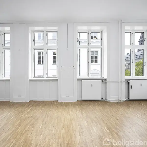 Et tomt rum med trægulv og tre store vinduer. Væggene er hvide, og der er radiatorer under vinduerne. Udenfor ses flere etager af en bygning og nogle træer.