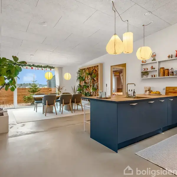 Køkken i moderne stil med blå køkkenø, hængelamper og indendørs planter, overgår til spisestue med stor spisebord og udsigt til terrasse gennem vinduer. Væghylder med dekorationer.