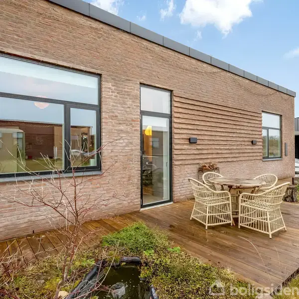 Et moderne rækkehus med murstensfacade har en terrasse med et bord og fire fletstole. Terrassen er omgivet af en lille havedam og græsplæne under en blå himmel.