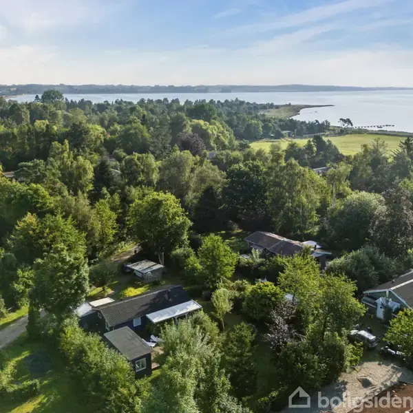 Huse omgivet af frodig skov ved kysten. Træer dækker området, mens havet breder sig i horisonten og skaber en fredfyldt atmosfære. Ingen tekst er i synlig i billedet.