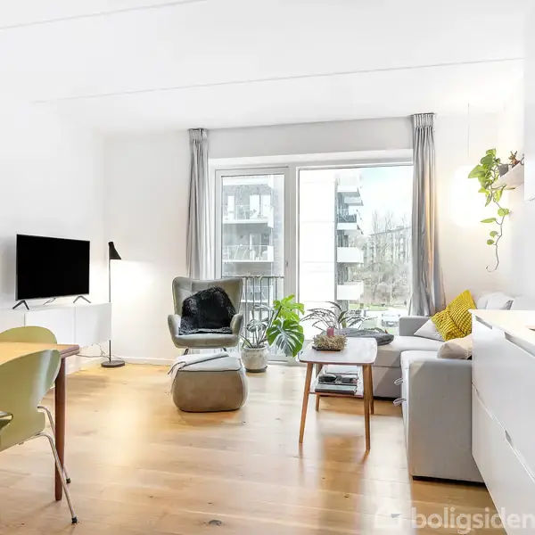 Stue med lyst trægulv har moderne møblering. Et spisebord med grønne stole står ved siden af et gråt sofaarrangement. Store vinduer lader naturligt lys skinne ind i rummet.