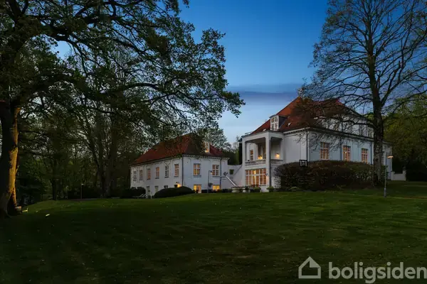 Stort hvidt hus med rødt tag står omgivet af tætte træer i en skumringsbelyst have. Husets lys er tændt, hvilket skaber en varm atmosfære mod den klare, blå aftenhimmel.