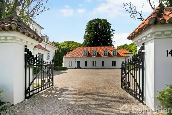 En hvid bygning med rødt tegltag står bag en tofløjet smedejernslåge. En grusindkørsel fører op til huset, omgivet af grønne træer. Nummeret "14" er synligt.