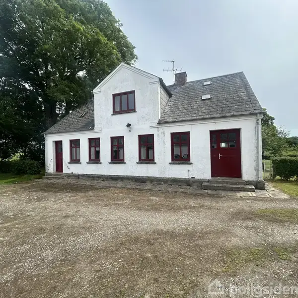 Et hvidt hus med røde vinduer og dør står i et landligt område. Der er en grusbelagt forplads, og store træer omkranser stedet.