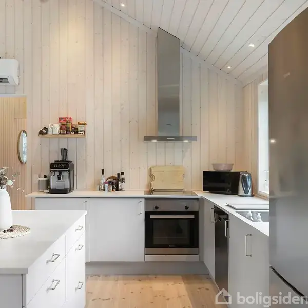 Et moderne køkken med hvidt træpanel på væggene har en ovn, emhætte, mikrobølgeovn og stort køleskab. En kaffemaskine og dekorationer findes på bordet. En airconditionenhed ses øverst.