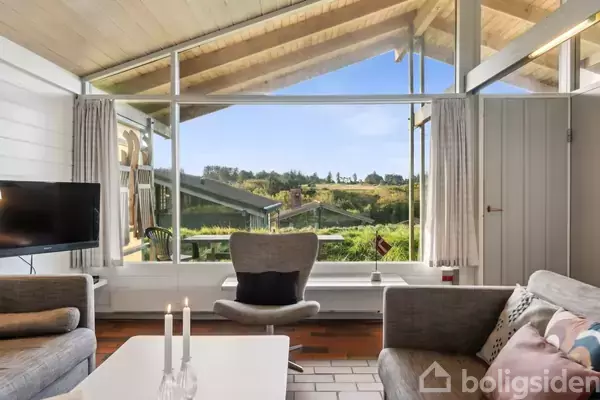 En stue med store glasvinduer giver udsigt til en grøn have og bakker. En sofa og stol er placeret omkring et sofabord med lysestager. En TV står ved siden af.