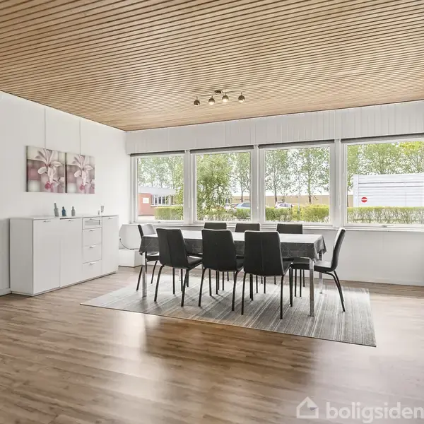 Spisebord med otte sorte stole står i et lyst rum med trægulv og træloft. Store vinduer giver udsigt til en grøn have. En hvid skænk står ved siden af.
