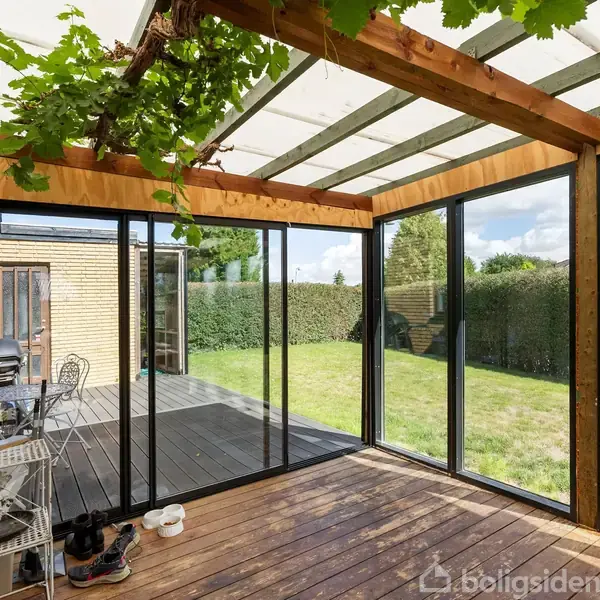 Overdækket terrasse med glastag og træramme, hvorfra vinstokke hænger. Terrasse fører til en have med græsplæne og hække. Dør åbner til en bygning med grill.
