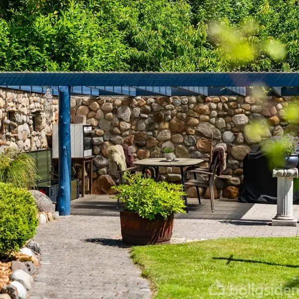En overdækket terrasse med et blåt trætag rummer et bord og stole. Omkring er der en stenmur og omgivende grønne planter, der skaber en hyggelig haveatmosfære.