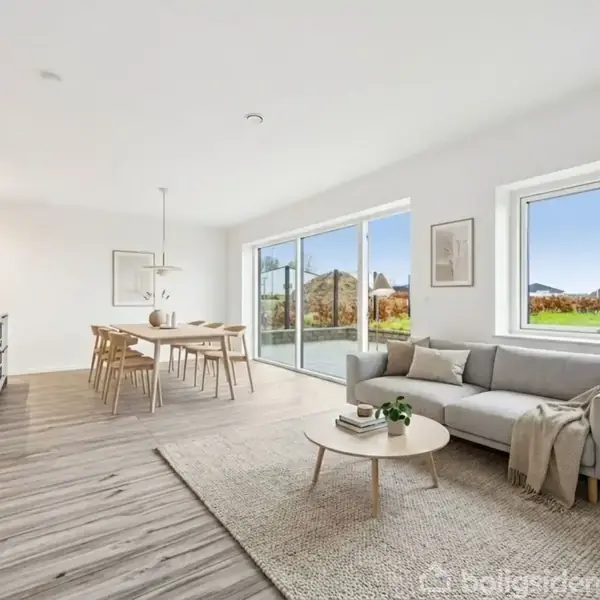 En lys stue med en grå sofa ved et vindue, et rundt sofabord, og en stueplante. Spisebord med stole står ved et køkken i baggrunden. Udsigt til natur udenfor.