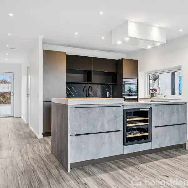 Køkkenø med grå skuffer og indbygget vinkøleskab står i et moderne, lyst køkken. Bagved ses høje sorte skabe og ovn, med en stor vindue til højre. Lang gang fører ud til en havedør.