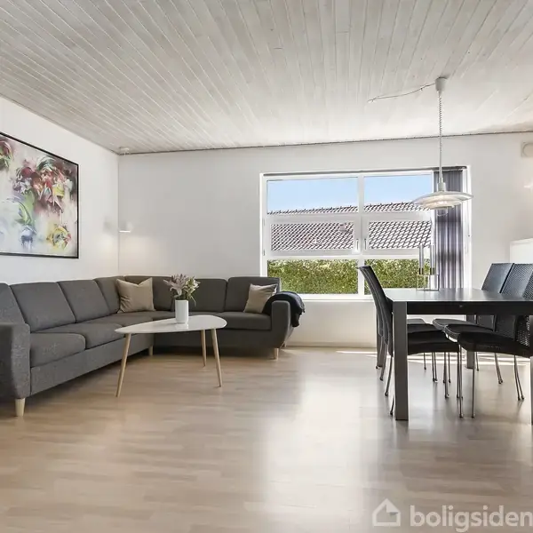 Brunt sofaarrangement står ved siden af et rundt sofabord i en stue, hvor et spisebord med stole er på højre side. Store vinduer giver udsigt til have og tag.