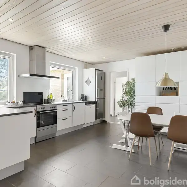 Et moderne køkken med hvide skabe og en grå bordplade. Køleskab og ovn står langs væggen. Et spisebord med brune stole er placeret til højre.