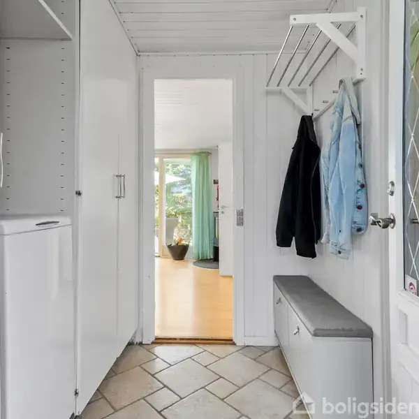Toilet står ved siden af håndvask i lyst badeværelse med hvide fliser. Bruseniche til venstre bag forhæng. Tre små, indrammede billeder på væggen. Tooth elementer på vasken.