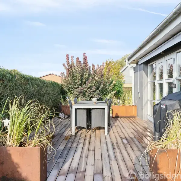 En terrasse med træbord står blandt rustikke plantekasser fyldt med grønt, ved siden af et hus med store vinduer. Baggrund består af en tæt hæk og klar blå himmel.