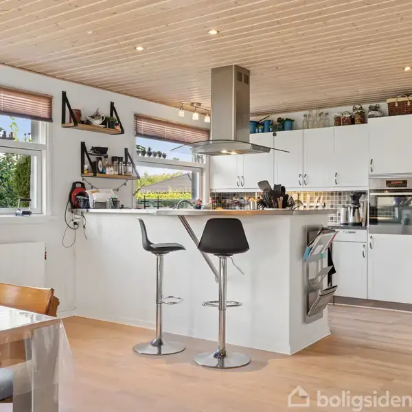 Et køkken med hvide skabe og en kogeø, hvor der står to sorte barstole. En grå emhætte hænger over kogeøen. Store vinduer giver udsigt til en have.