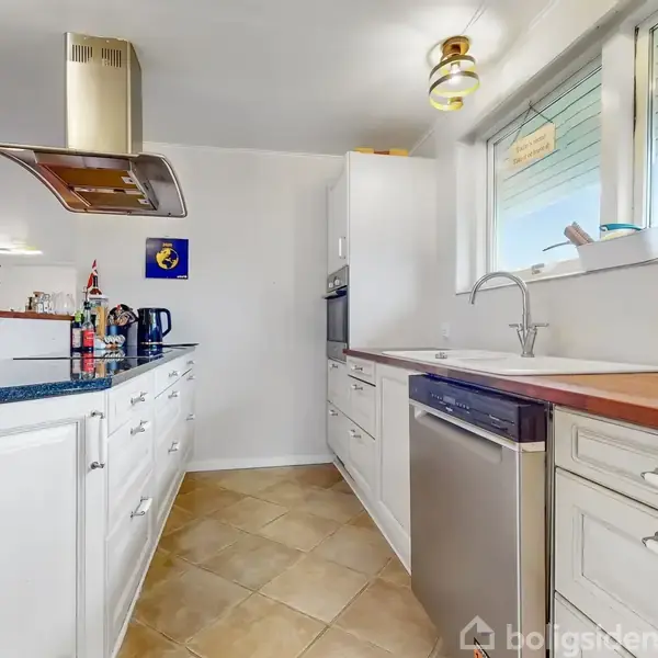 Et lyst køkken med hvide skabe og stålgreb, hvor en vask og opvaskemaskine er placeret under et vindue. Kanden står på bordet, og lys hænger fra loftet.