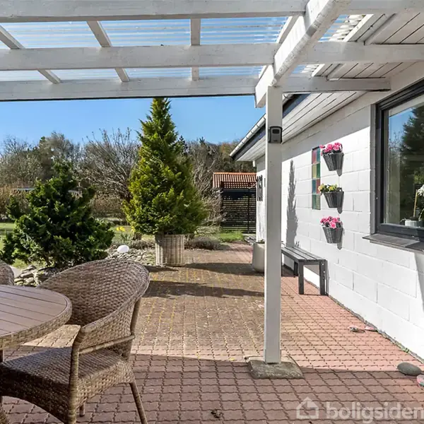 Rundt bord med fletstole står på en flisebelagt terrasse under en hvid pergola. Ved siden af er der en mur med blomsterkasser, og der ses en have med træer i baggrunden.