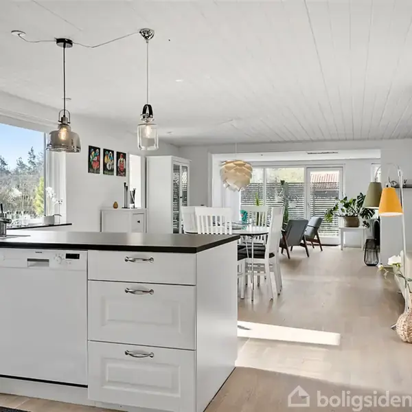 Et lyst køkken med hvide skabe og sort bordplade. Spisebord med stole i baggrunden. Store vinduer giver udsigt til træer udenfor. Gulvlampe og planter står langs væggen.