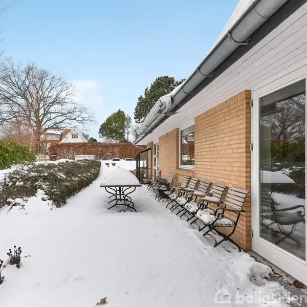 En murstensbygning står ved siden af en snedækket gårdhave, hvor flere stole og et bord er placeret. Træer og buske omkranser området, og himlen er klar blå.