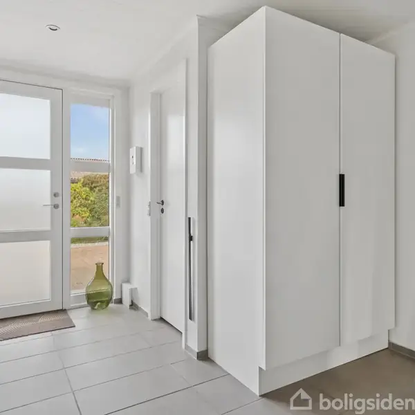 En hvid garderobe står stille i en lys entré. Frakke med pelskrave hænger på en knage, og en grøn vase står ved vinduet med udsigt til en have.