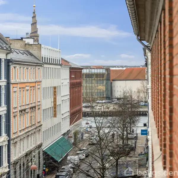 Bygninger står langs en gade, hvor folk går og biler kører. I baggrunden ses et højhus og tårnet på en historisk bygning. Himlen er klar og blå.