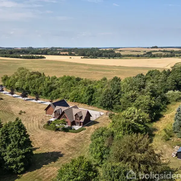 Et rødstenshus ligger omgivet af grønne træer på en stor landlig grund. Husets tag er mørkt, og der ses en grusvej samt marker og skove i baggrunden.