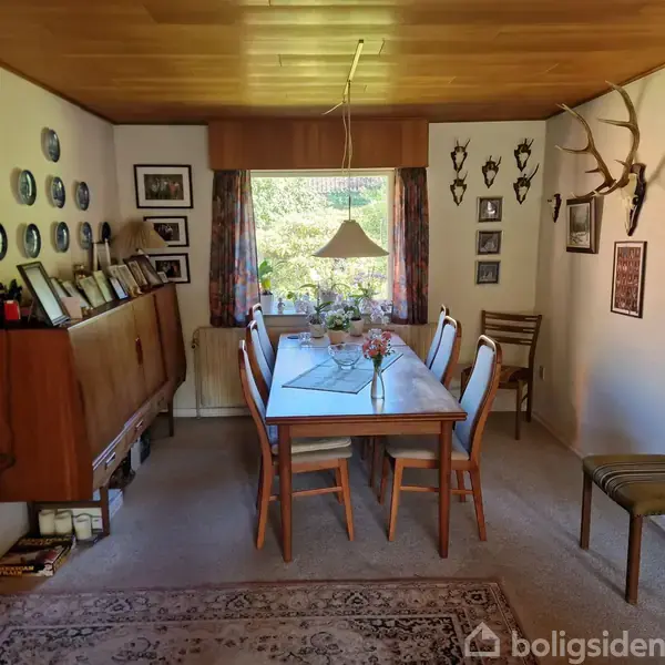 Et spisebord er dækket med en vase og dekorative genstande, omgivet af stole. I baggrunden ses en stor vindue med gardiner, en kommode med billeder, og vægge udsmykket med tallerkener og gevirer.