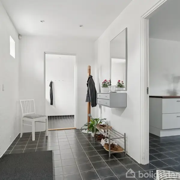 En gang med sort flisegulv og hvidmalede vægge. Et spejl hænger over en moderne dekorationshylde. En hvid stol står til venstre, mens en stueplante står på gulvet.