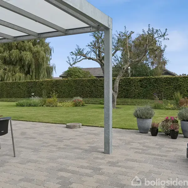 Overdækket terrasse med borde og stole står på flisebelagt område. I baggrunden ses en velplejet have med græsplæne, buske og træer mod en blå himmel.