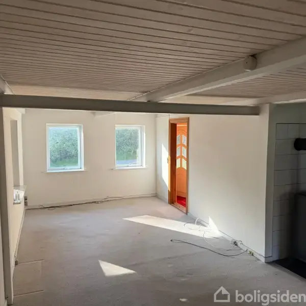 Et tomt rum med hvide vægge og loftsbjælker. En brændeovn står til højre, mens to vinduer bader rummet i lys. En åben dør fører til en anden sektion.