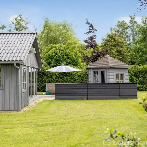 Et gråt træhus står stille på en velplejet græsplæne. I baggrunden ses en mindre pavillon i samme stil, omgivet af tætte grønne hække og høje træer.